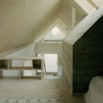 Podkrovná spálňa s prírodným dreveným obkladom, nízkym policovým regálom a hrubou betónovou stenou.