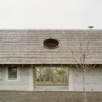 Fasáda domu z hrubozrnnej omietky s tmavou šindľovou krytinou a výrazným kruhovým otvorom v streche.