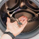 Detail na ruku držiacu kľúče a sponku do vlasov pred otvoreným bubnom práčky.