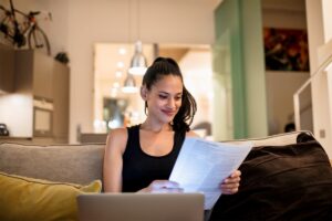 Žena sedí v obývačke na sedačke pri notebooku s dokumentmi v ruke.