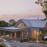 Moderný rodinný dom so sivou plechovou strechou, kamenným obkladom a veľkými presklenými plochami v lesnom prostredí.