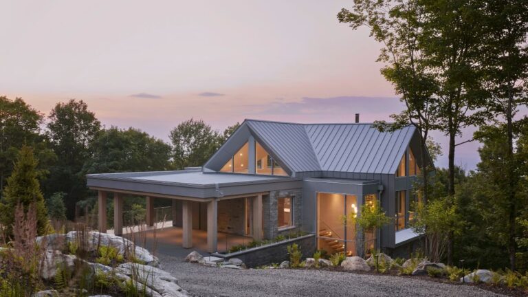 Moderný rodinný dom so sivou plechovou strechou, kamenným obkladom a veľkými presklenými plochami v lesnom prostredí.