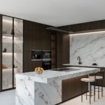 Minimalistická kuchyňa s tmavým dreveným dekorom, mramorovým obkladom a bielym ostrovom s barovými stoličkami.