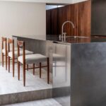 Kuchyňa s nerezovým ostrovom, drevenými dizajnovými stoličkami a svetlou travertínovou podlahou.