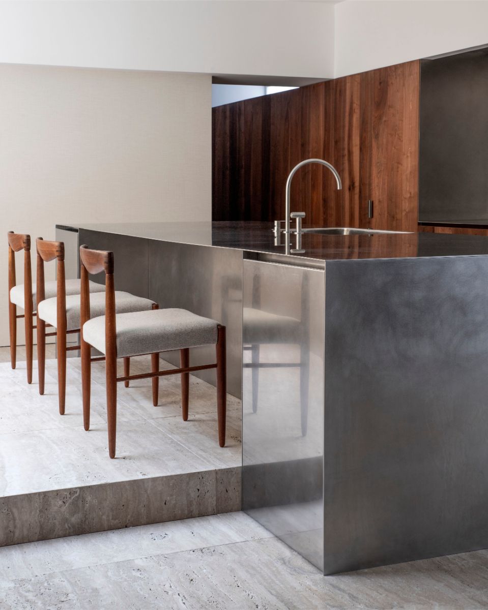Kuchyňa s nerezovým ostrovom, drevenými dizajnovými stoličkami a svetlou travertínovou podlahou.