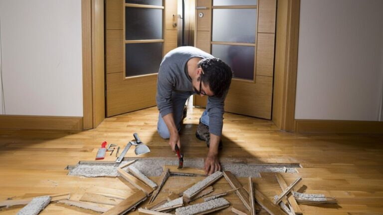 Kľačiaci muž s kladivom a sekáčom pri oprave drevenej parketovej podlahy.