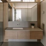 Kúpeľňa s drevenou plávajúcou skrinkou, sivou pracovnou doskou a veľkým trojdielnym zrkadlom.