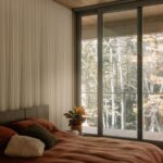 Spálňa s hnedou posteľnou bielizňou, presklenou stenou a výhľadom na jesenný listnatý les.