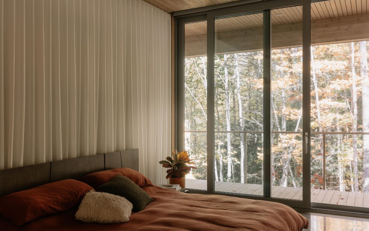 Spálňa s hnedou posteľnou bielizňou, presklenou stenou a výhľadom na jesenný listnatý les.