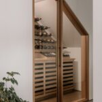 Chodba s presklenou vinotékou pod schodmi, drevenými policami a čiernymi kovovými úchytmi.