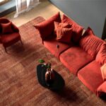 Oranžová zamatová sedačka s olivovou dekou, drevený stolík a krémová stena s čiernymi rámami obrazov.