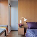 Spálňa s dreveným stenovým panelom, malým samonosným stolíkom, bielou polohovateľnou lampou a fialovými vankúšmi.