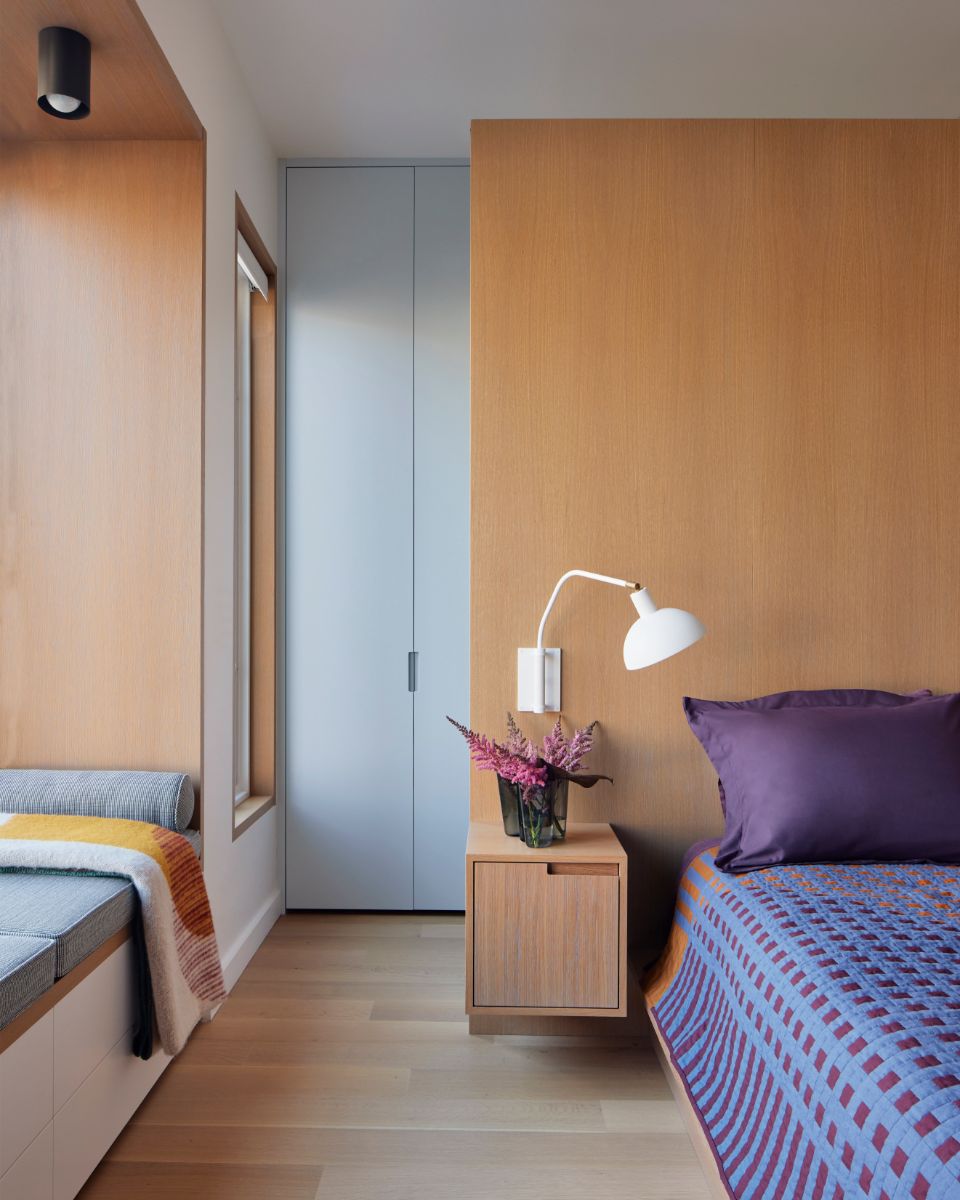 Spálňa s dreveným stenovým panelom, malým samonosným stolíkom, bielou polohovateľnou lampou a fialovými vankúšmi.