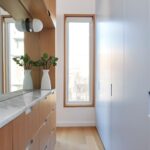 Šatník so svetlými vstavanými skriňami, drevenou komodou, mramorovou doskou a vysokým oknom.