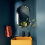 Toaleta s tmavomodrými stenami, žltým betónovým umývadlom a oválnym zrkadlom s čiernym rámom.