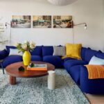 Obývacia izba s výraznou modrou sedačkou, okrúhlym dreveným stolom a zeleným tkaným kobercom.