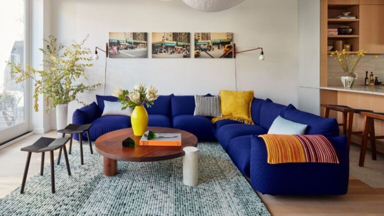 Obývacia izba s výraznou modrou sedačkou, okrúhlym dreveným stolom a zeleným tkaným kobercom.