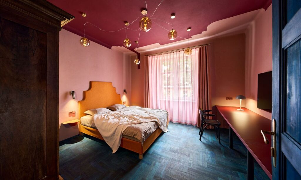 Spálňa s oranžovou posteľou, vínovočerveným stropom, modrou drevenou podlahou a visiaci sklenenými svietidlami.