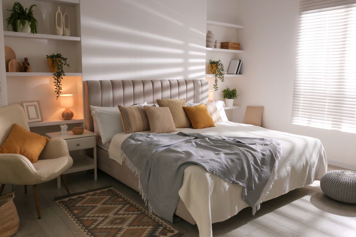 Svetlá spálňa s čalúnenou posteľou, horčicovými vankúšmi, bielymi vstavanými policami a vlneným krémovým kreslom.