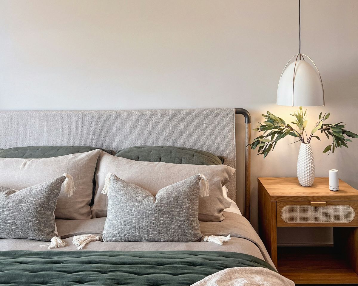 Detail spálne s čalúneným čelom postele, zelenými vankúšmi, dreveným nočným stolíkom a bielym visiacim svietidlom.