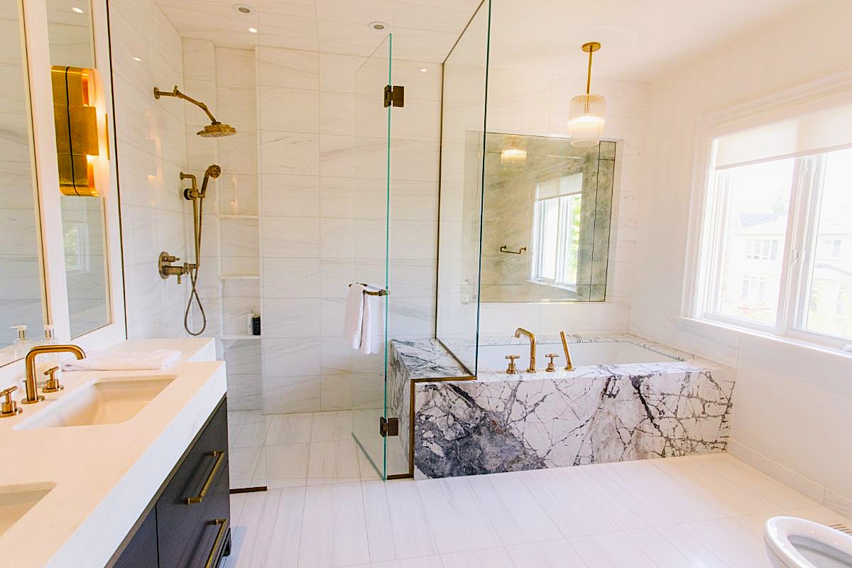 Moderná biela kúpeľňa s mramorovou vaňou, walk in sprchovým kútom a zlatými armatúrami.