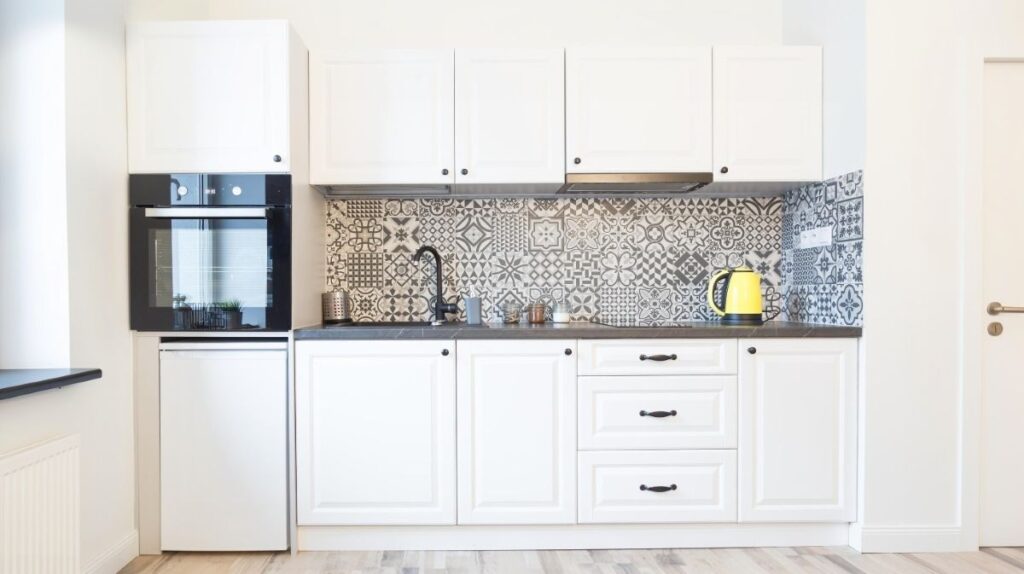 Škandinávska biela kuchyňa s tmavou doskou, čiernym kohútikom, vzorovaným obkladom, vstavanou rúrou a žltou kanvicou.