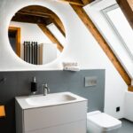Kúpeľňa v podkroví s bielou sanitou, podsvieteným zrkadlom a šedým obkladom.