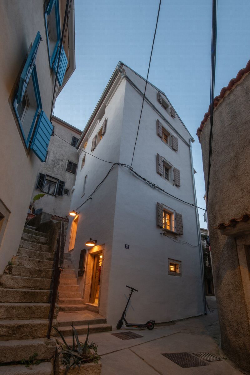 Viacposchodový biely dom s drevenými okenicami, kamennými schodmi a úzkou uličkou za súmraku.
