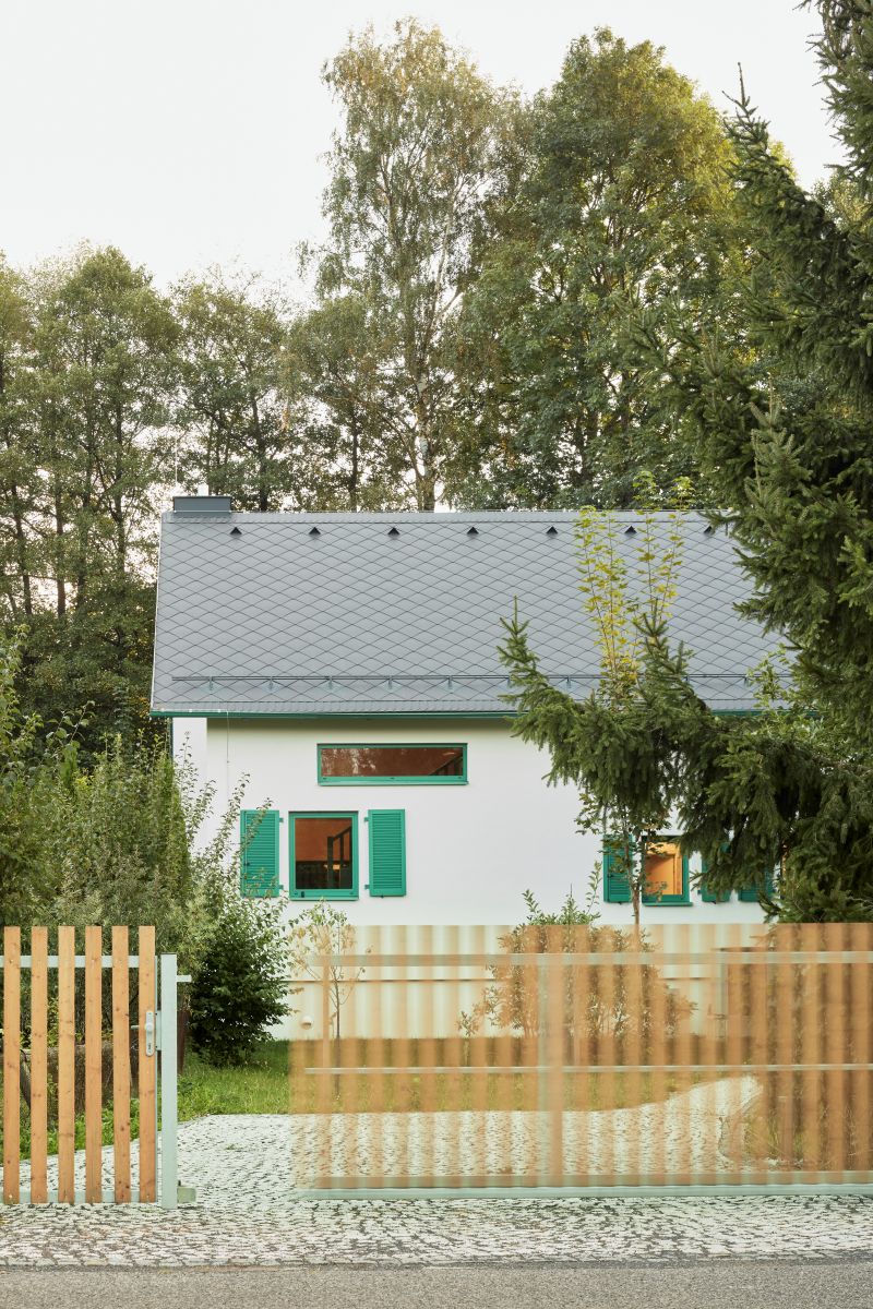 Drevený latkový plot pred bielym domom so sivou strechou, zelenými rámami okien a dláždeným chodníkom z mačacích hláv.