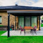 Čierna hliníková pergola nad šedou kamennou terasou s tehlovým obkladom domu a dreveným jedálenským stolom.