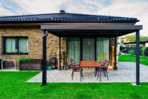 Čierna hliníková pergola nad šedou kamennou terasou s tehlovým obkladom domu a dreveným jedálenským stolom.
