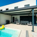 Moderná antracitová pergola pri bielom rodinnom dome s bazénom, sivou pohovkou a čiernymi vonkajšími žalúziami.