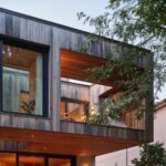 Moderná architektúra s drevenou fasádou, zapustenou hornou terasou, veľkými oknami a listnatým stromom v popredí.
