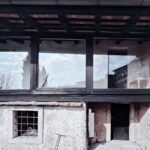 Detail omietnutej steny s mrežovaným oknom, čiernymi dverami a starou kovovou kolobežkou na štrku.