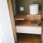 Drevený nočný stolík s bielou zásuvkou, stolovou lampou a zelenou štruktúrovanou tapetou.