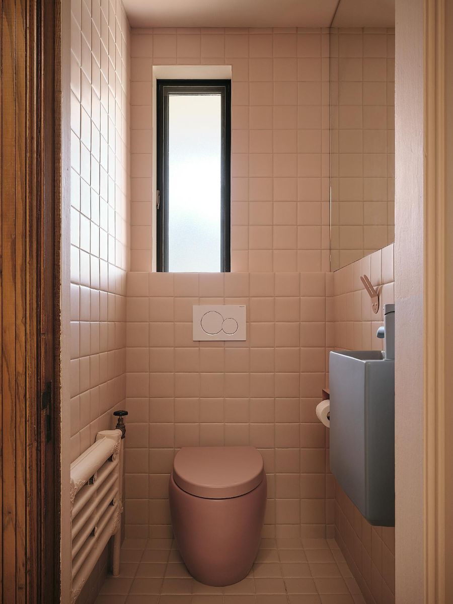 Toaleta s ružovým obkladom, závesným ružovým wc, sivým minimalistickým umývadlom a úzkym oknom.